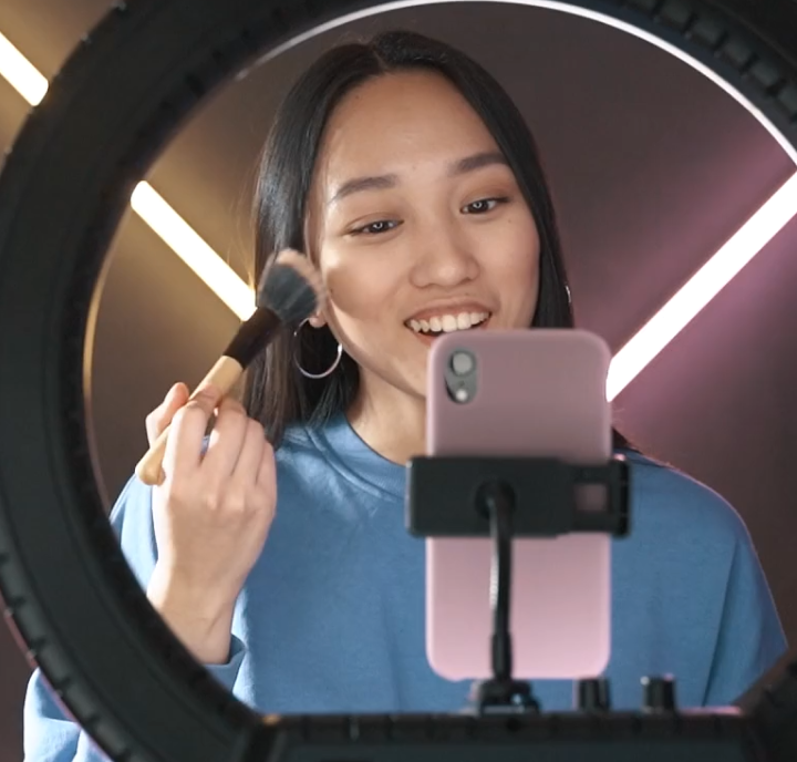 A woman in a blue sweater applies makeup in front of a ring light and phone.